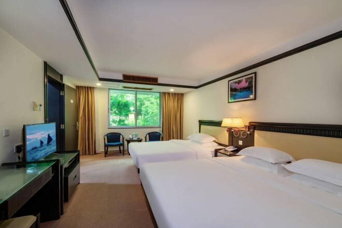 A hotel room with two beds, a desk with a television, a small table with chairs, and a window with curtains.