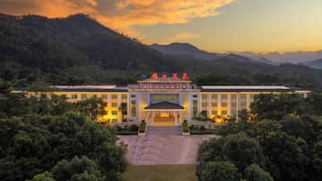 Un hotel grande con entrada central, rodeado de abundante vegetación y montañas al fondo.