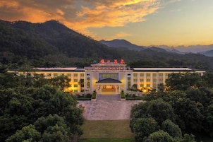 Un hotel grande con entrada central, rodeado de abundante vegetación y montañas al fondo.