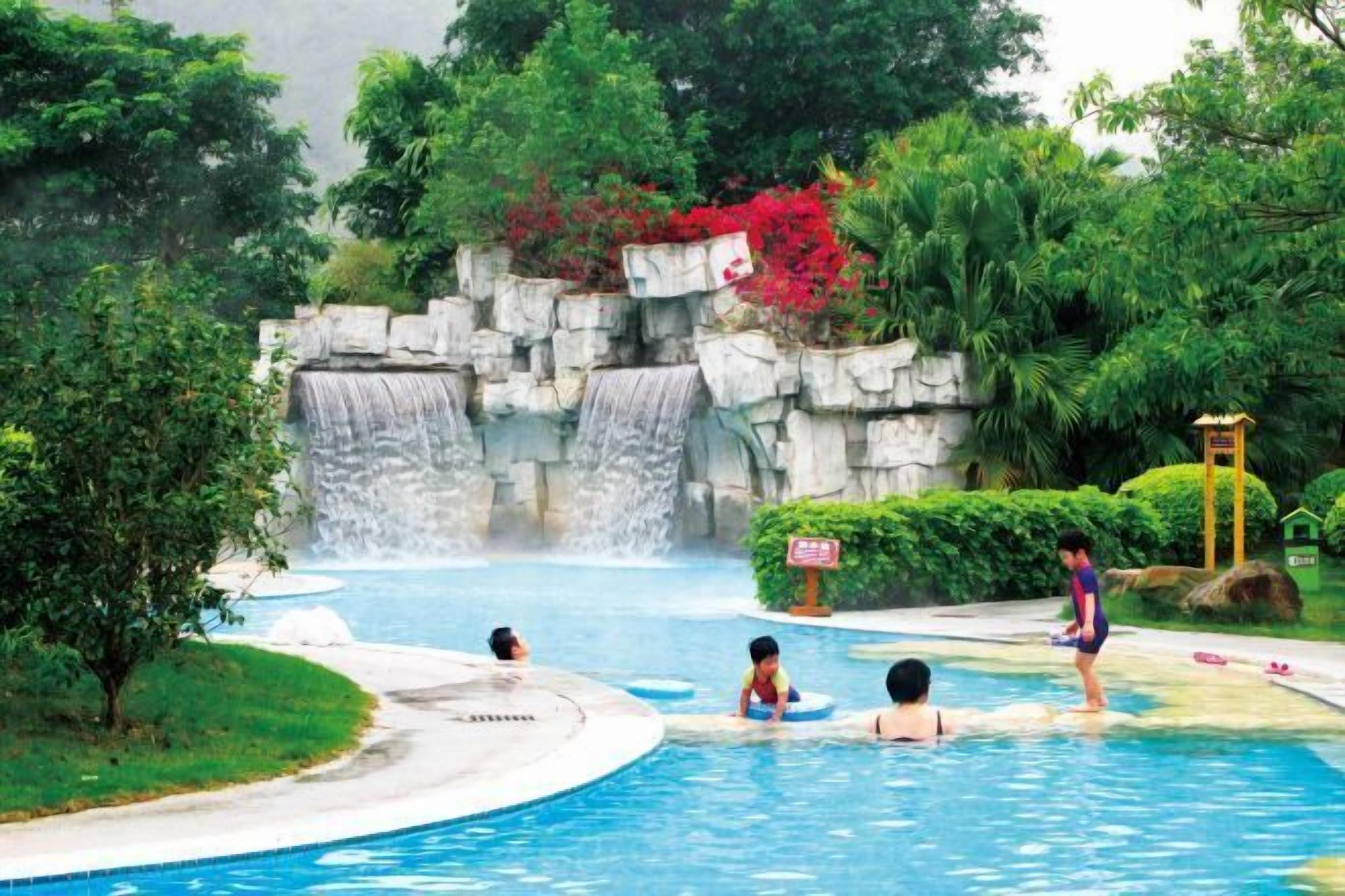 A swimming pool with a waterfall feature, surrounded by lush greenery and a stone wall.
