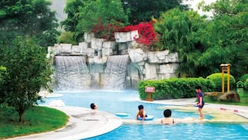 Una piscina con una cascada, rodeada de vegetación exuberante y un muro de piedra.