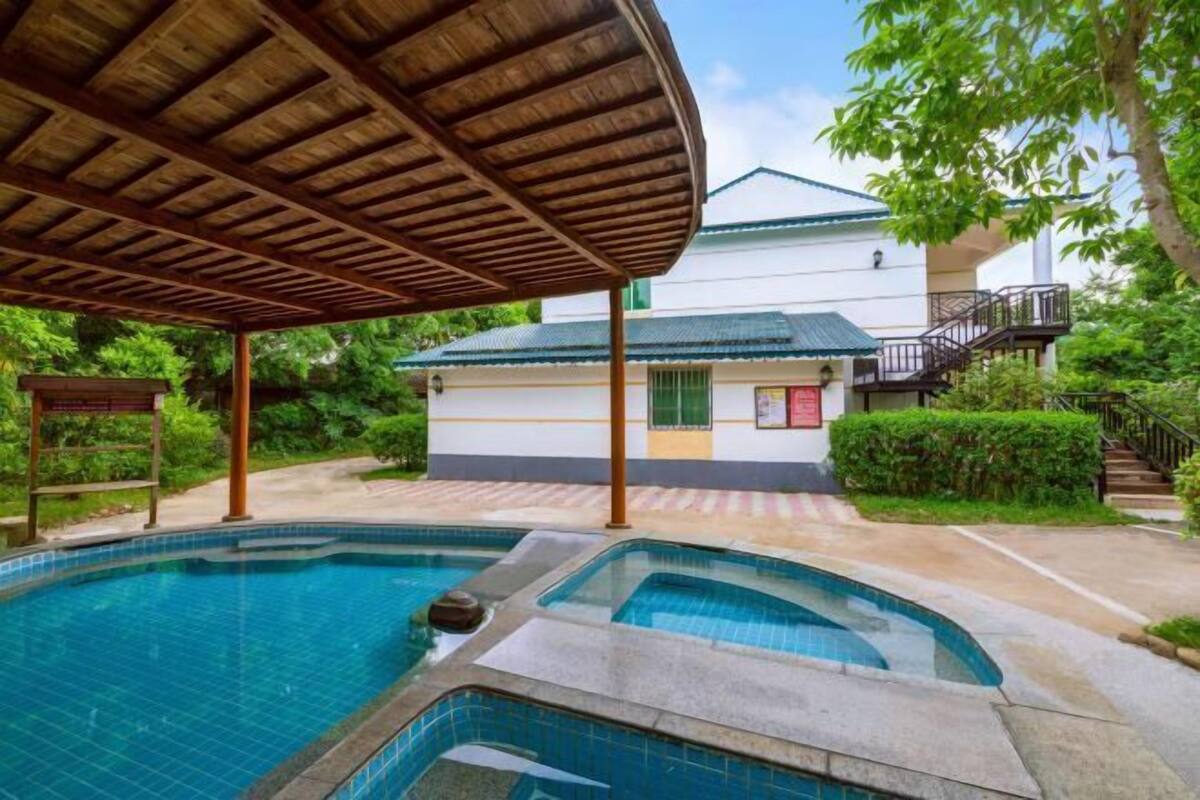 A swimming pool with a wooden canopy, a white building with a green roof, and a staircase leading to a balcony.