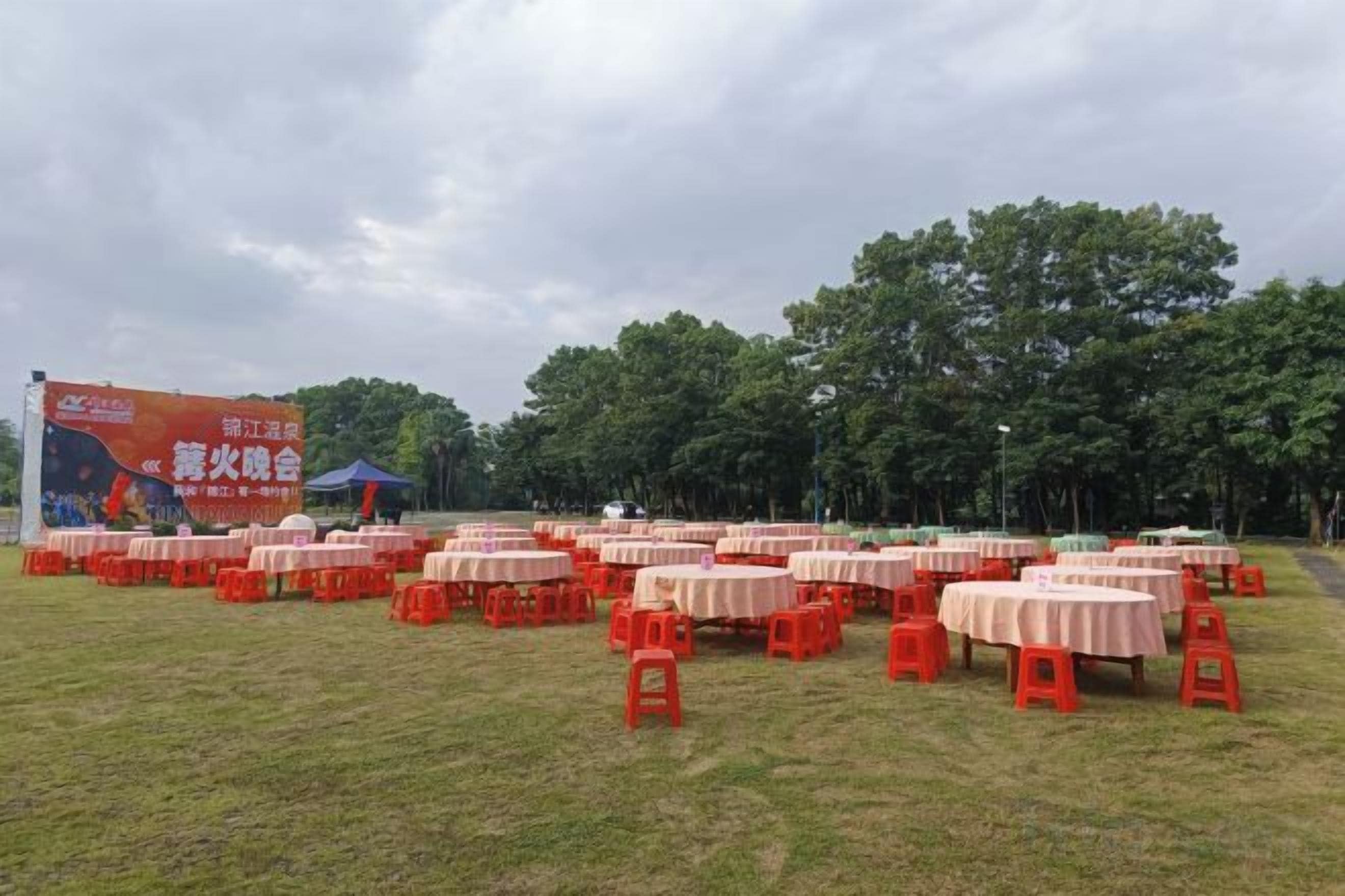 Un espacio para eventos al aire libre con varias mesas redondas y sillas, un cartel grande y árboles de fondo.