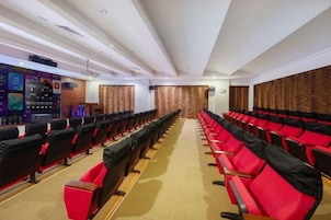 Un salón de conferencias con hileras de sillas negras y rojas, un escenario con un atril y piso alfombrado.
