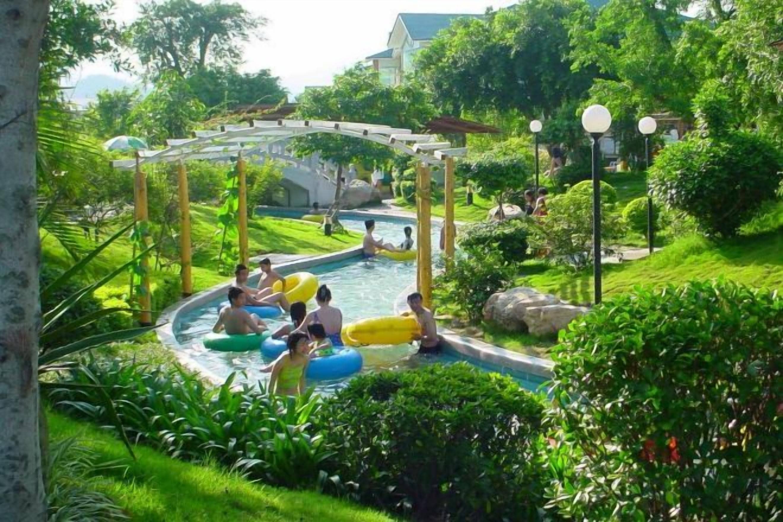 A swimming pool area with a wooden structure overhead, surrounded by lush greenery.