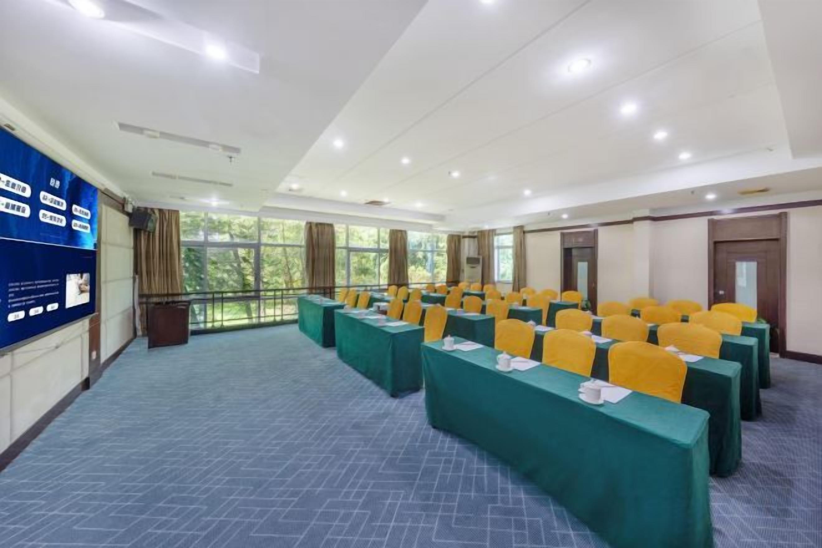 Un salón de conferencias con filas de mesas y sillas, ventanas amplias y una pantalla para presentaciones.
