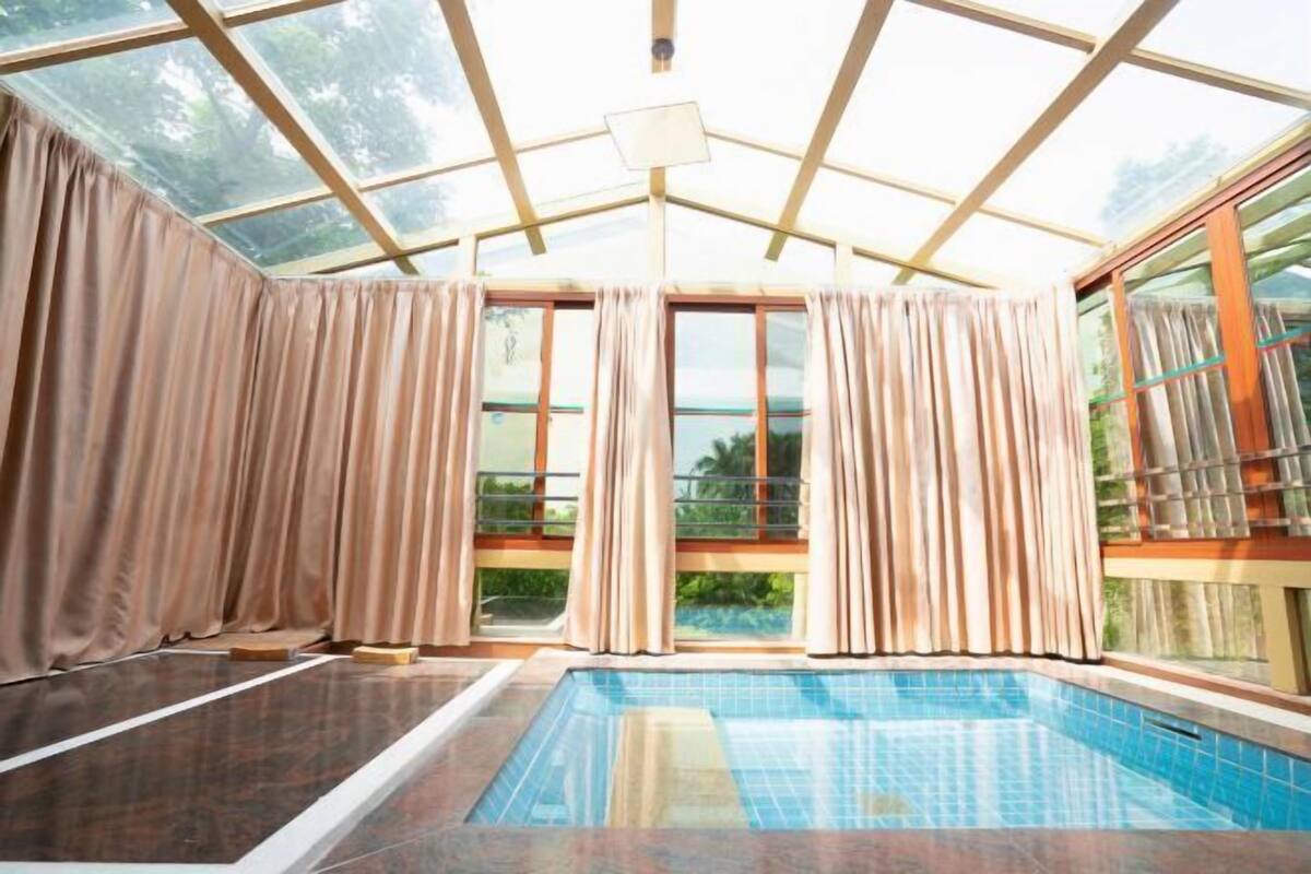 A swimming pool with a glass ceiling, beige curtains, and a view of greenery outside.