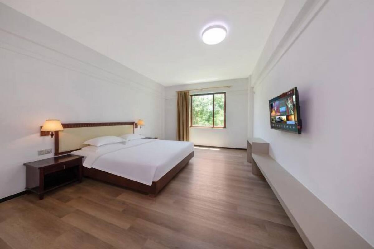 A hotel room with a wooden floor, a bed with white linens, a nightstand with a lamp, a window with curtains, and a flat-screen TV mounted on the wall.
