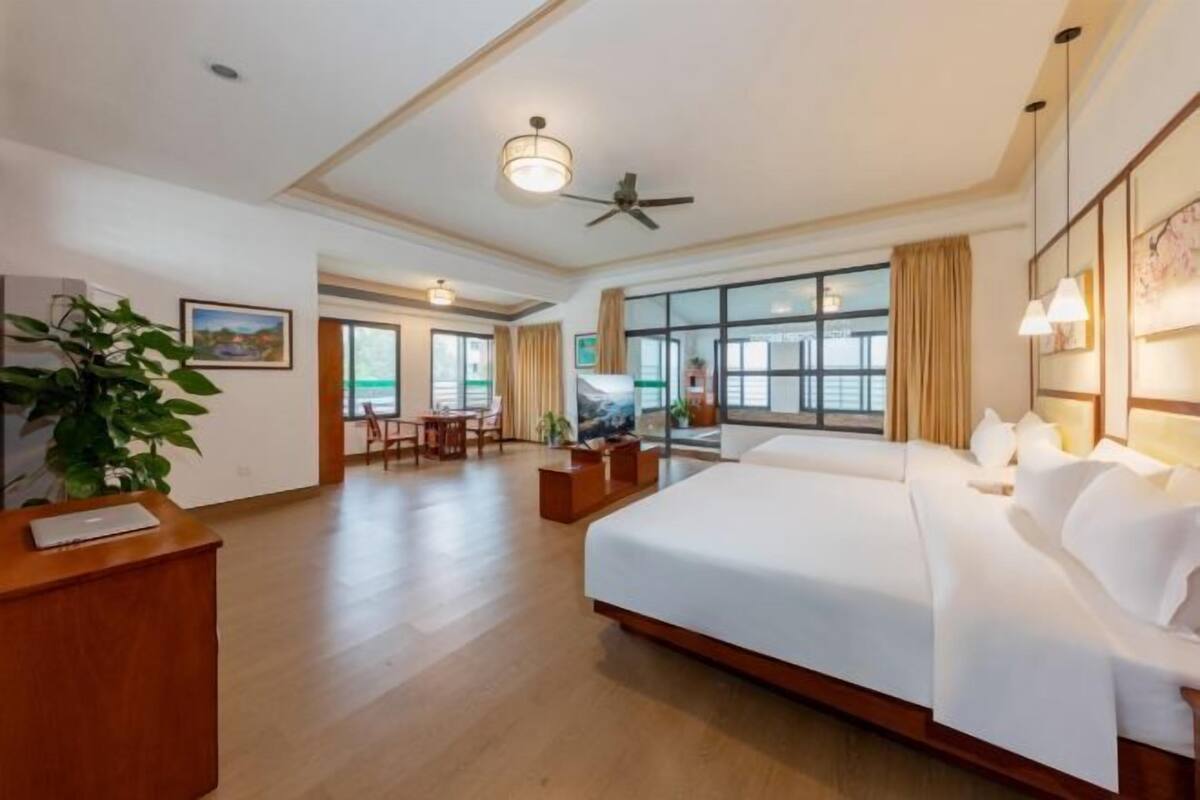 A hotel room with a large bed, a desk, a chair, a ceiling fan, and large windows.