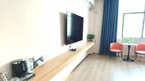 A hotel room with a flat-screen TV mounted on the wall, a wooden console table with a coffee maker and glasses, two red chairs around a small table, and a potted plant. - Jinse Yangguang Motel (Kuqa)