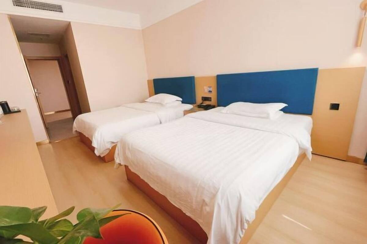 Habitación de hotel con dos camas, piso de madera, cabecera azul y una mesita de noche pequeña con lámpara.