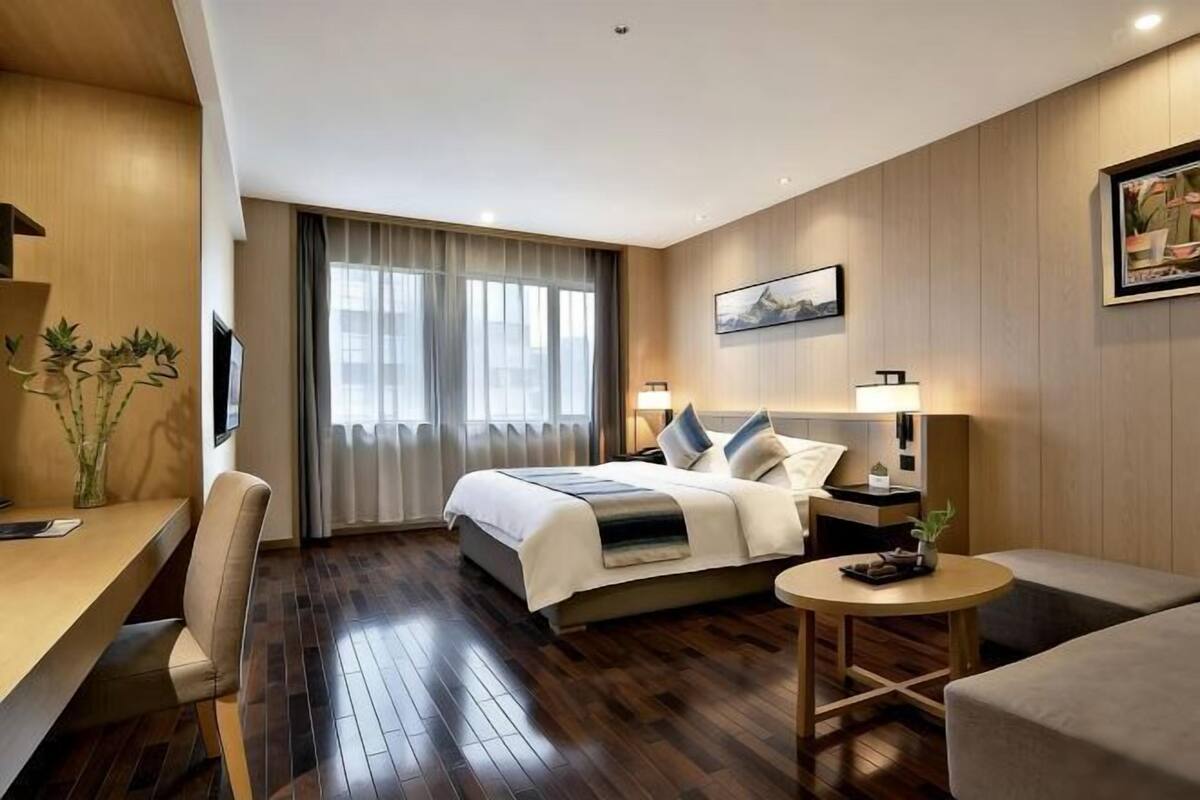 A modern hotel room with a bed, a desk, a chair, a round table, and a large window with curtains.