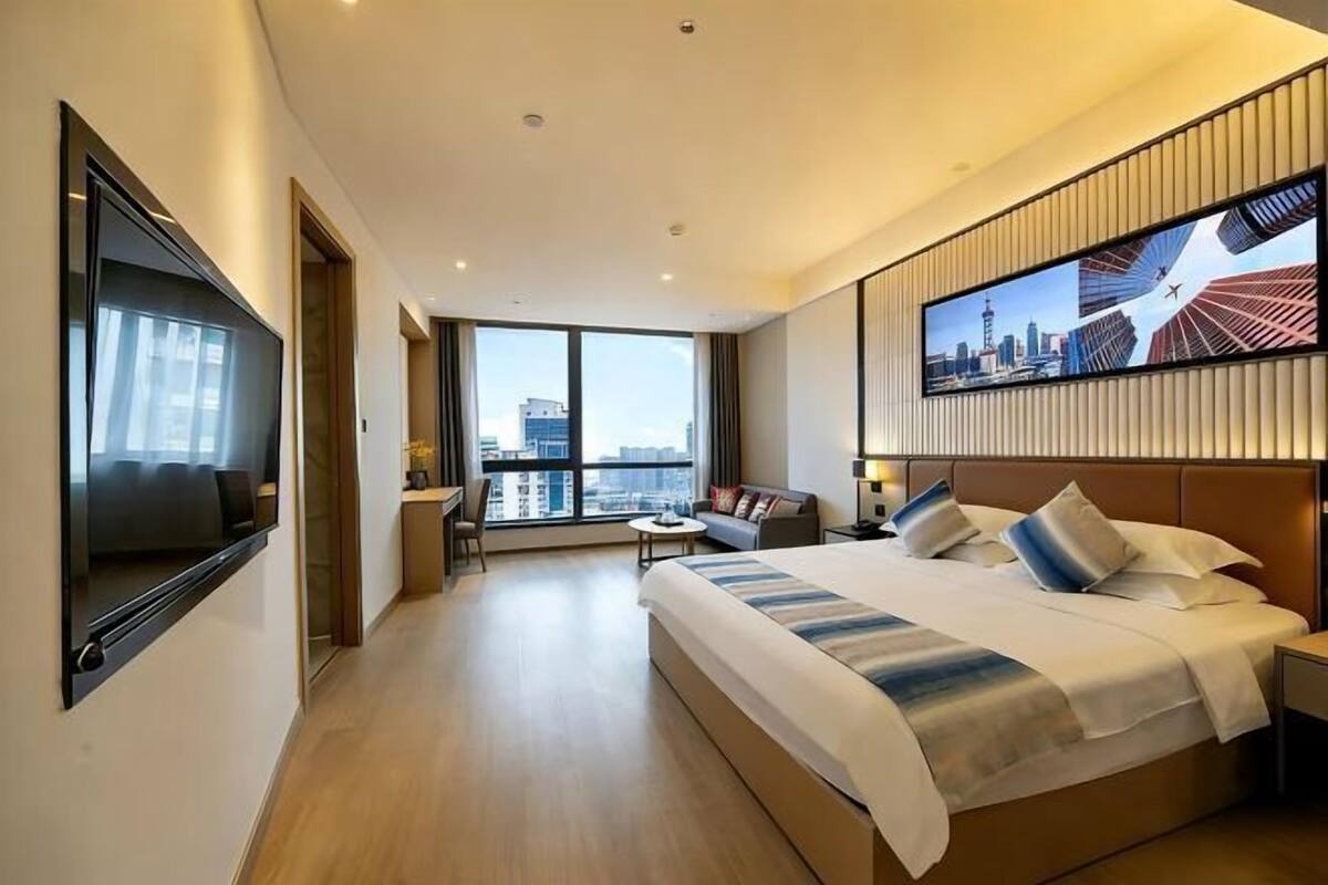 A modern hotel room with a large bed, a TV mounted on the wall, a seating area with a sofa and chairs, and a cityscape view through the window.