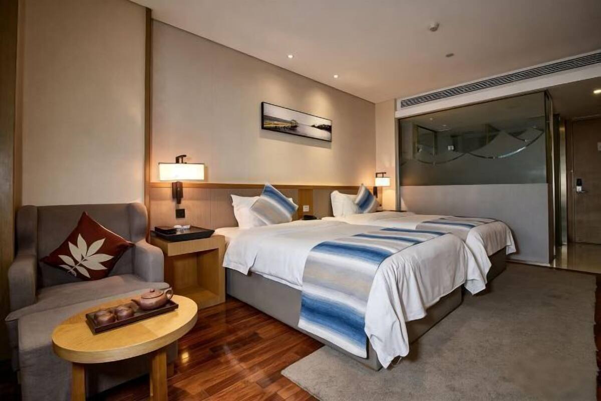 A hotel room with two beds, a sofa, a small table with a tray of teapots, a painting on the wall, and a sliding door.