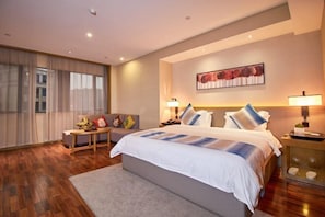A modern hotel room with a large bed, a sitting area with a sofa and armchairs, a large window with sheer curtains, and a painting on the wall.