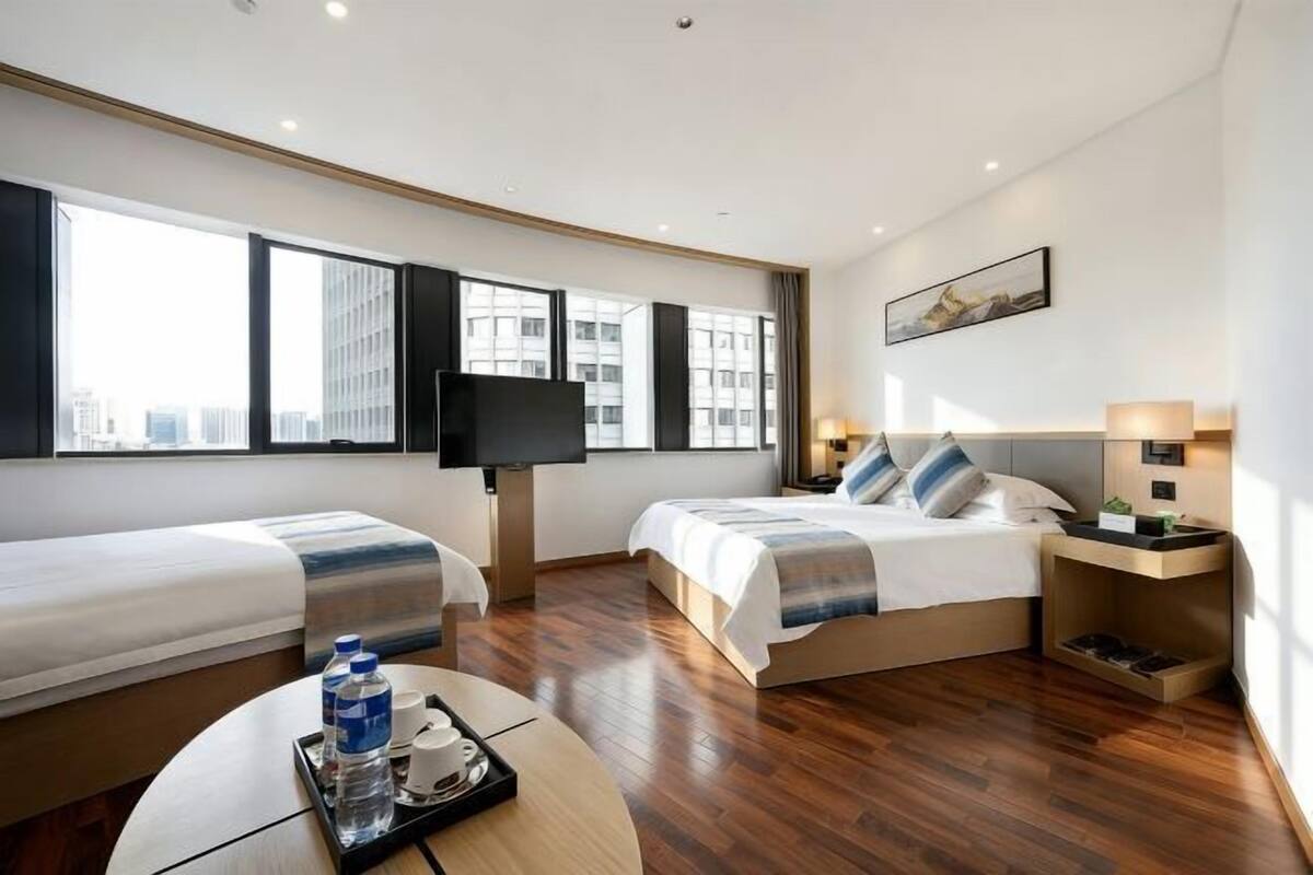 A hotel room with two beds, a TV, a desk with a tray of water bottles, and a view of the city.