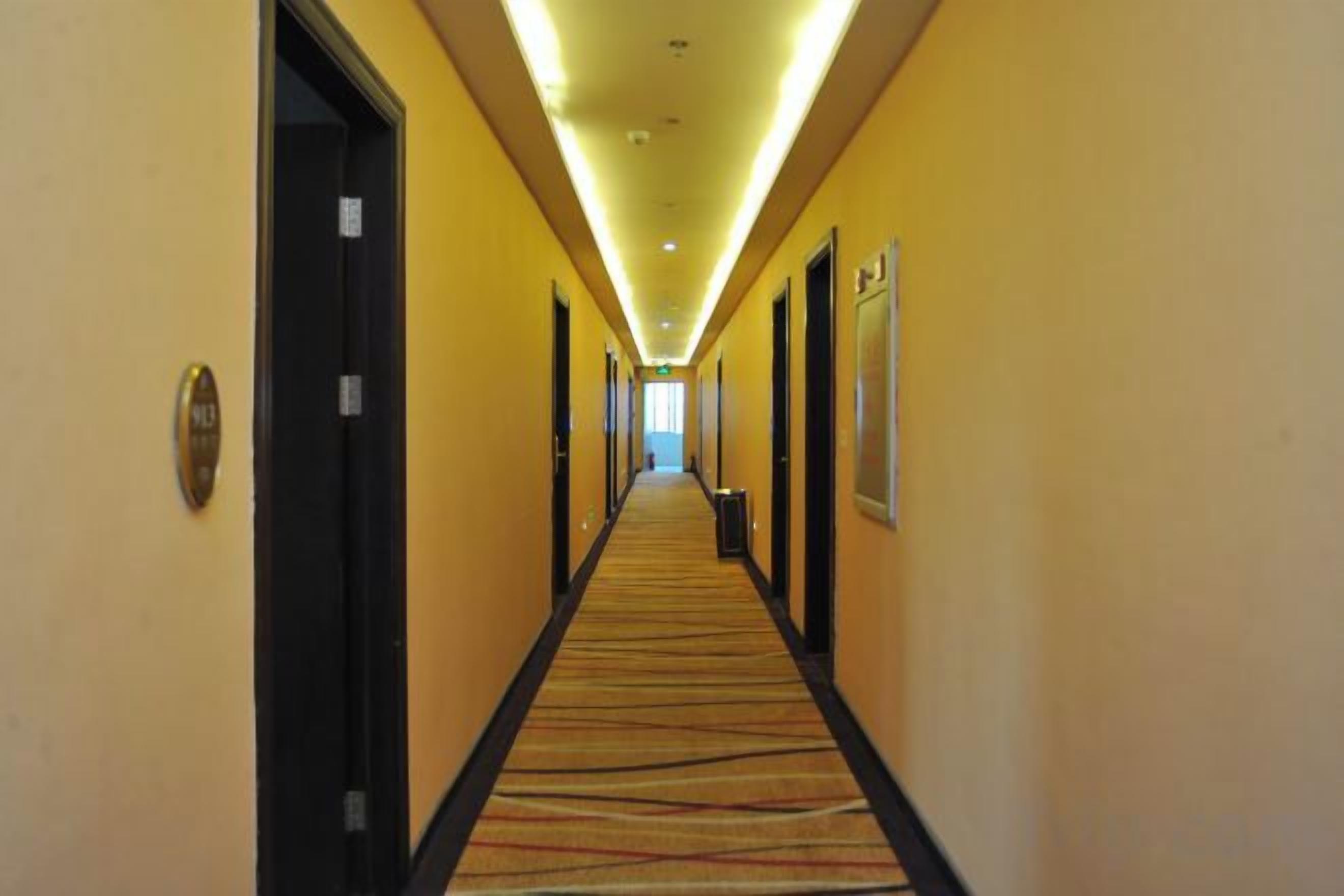 Un pasillo de hotel con puertas a ambos lados, piso alfombrado y un largo corredor que conduce a una zona luminosa al final.