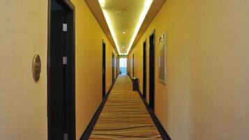 Un pasillo de hotel con puertas a ambos lados, piso alfombrado y un largo corredor que conduce a una zona luminosa al final.