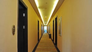 Un couloir d’hôtel avec des sols recouverts de moquette, des portes de part et d’autre, et un long couloir menant à une sortie lumineuse.