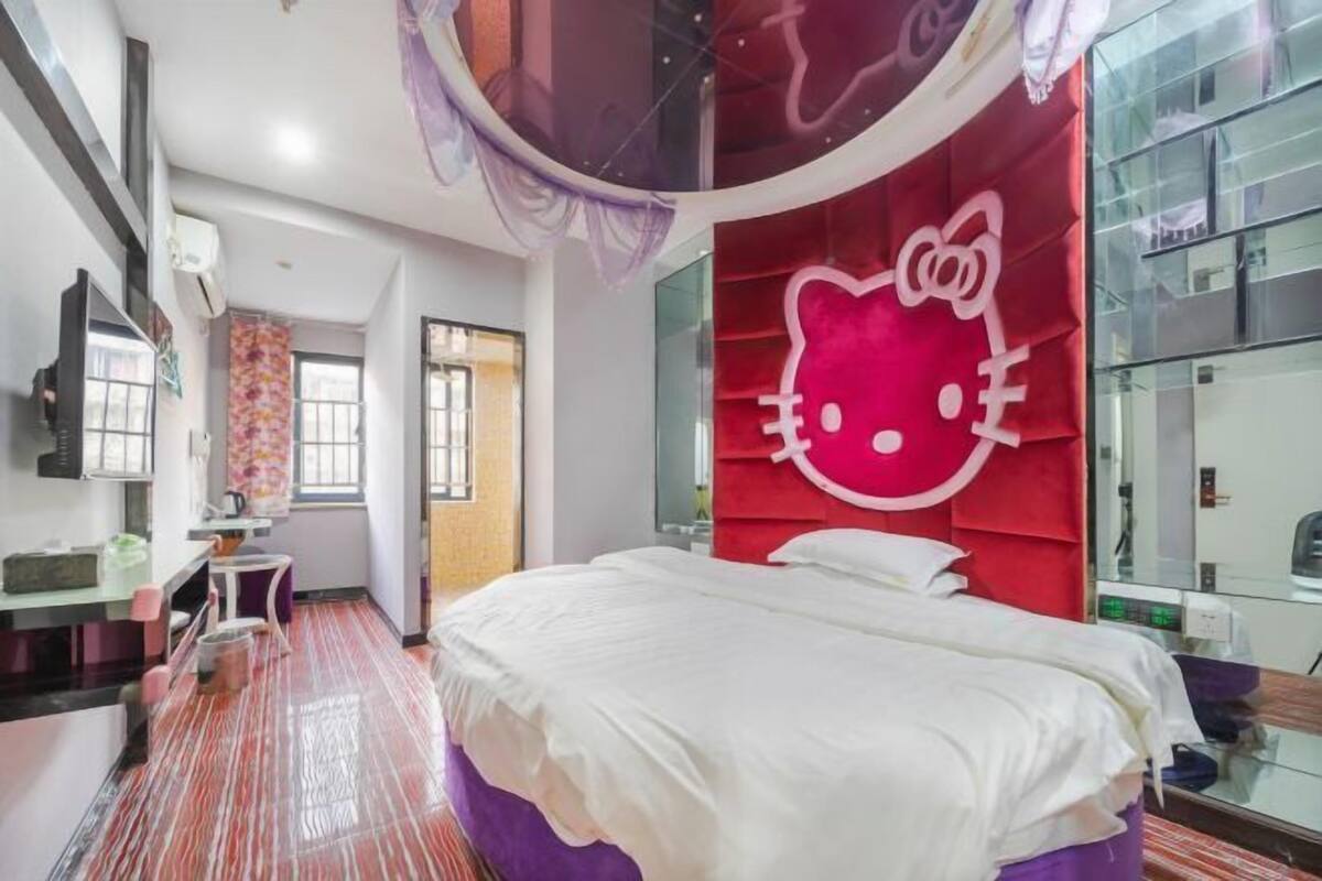 A modern hotel room with a large bed, a vanity with a mirror, and a red wall with a Hello Kitty design.
