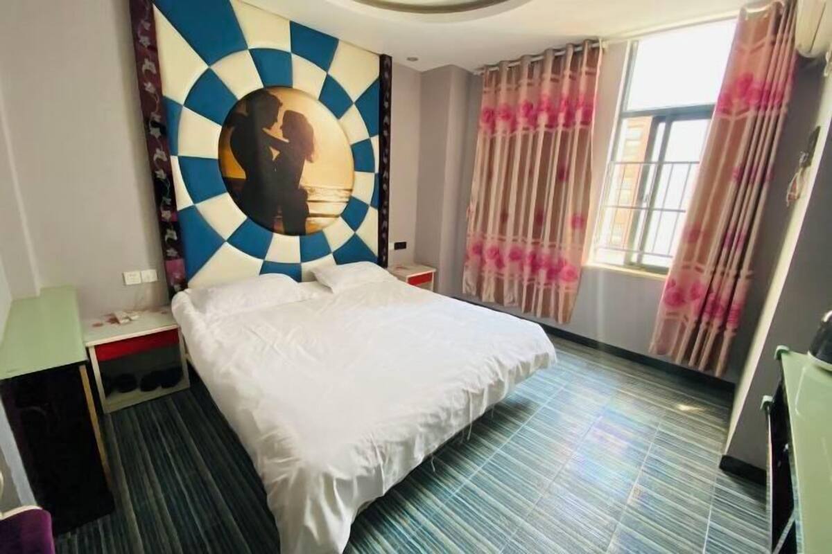 A hotel room with a bed, a nightstand, a wall art piece, a window with curtains, and a striped carpet.