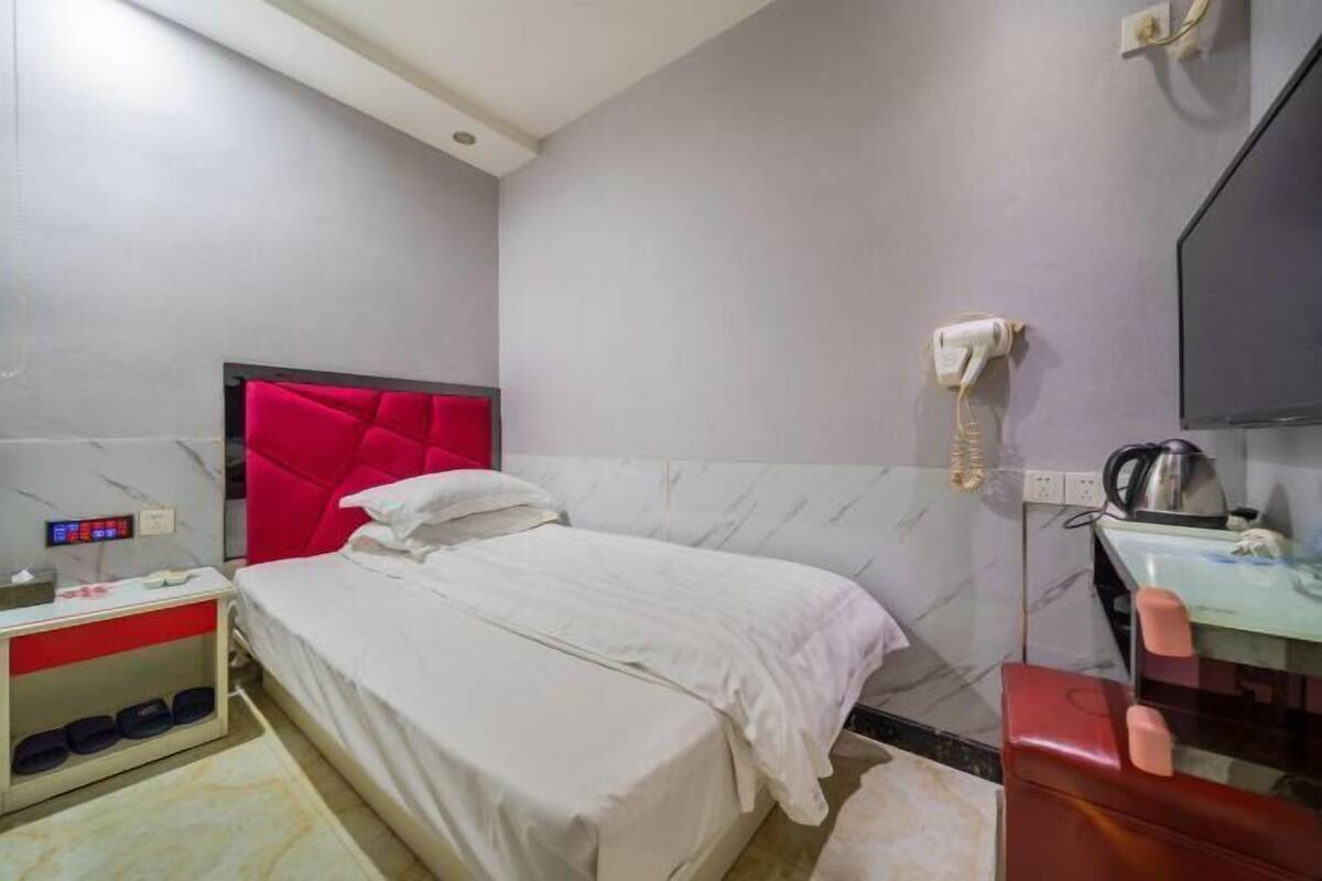A hotel room with a bed, a red headboard, a bedside table with a lamp, a mirror, and a wall-mounted phone.