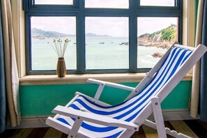 A room with a window view of a coastal area, a chair, and a vase with flowers.