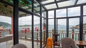 A glass-enclosed terrace with tables and chairs, overlooking a coastal area with boats and cliffs. - Haiyi Jiage Inn (Shengsi)