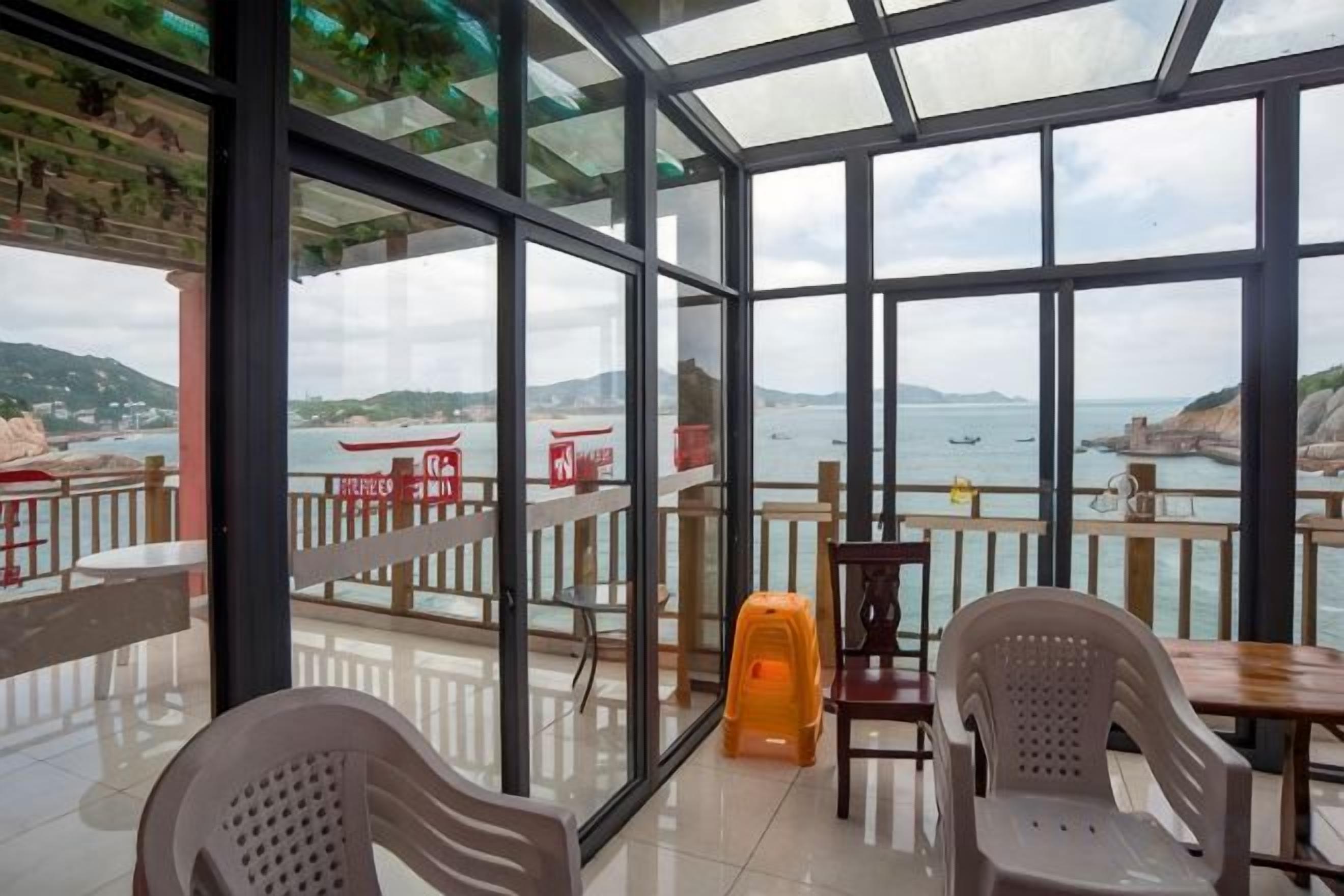 A glass-enclosed terrace with tables and chairs, overlooking a coastal area with boats and cliffs.