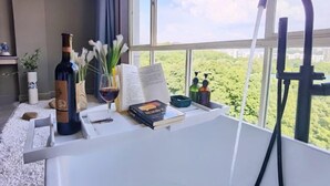 Een kamer met een raam, een badkuip, een fles, een boek en een vaas met bloemen.