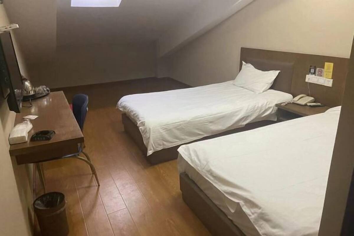 A small, single-room hotel room with two single beds, a desk with a chair, a trash bin, and a small window.