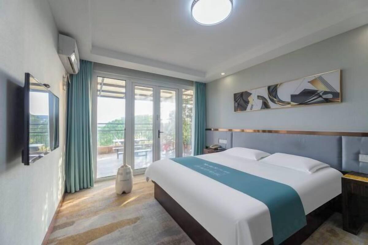 A hotel room with a bed, a television, a painting, and a view of the outdoors.