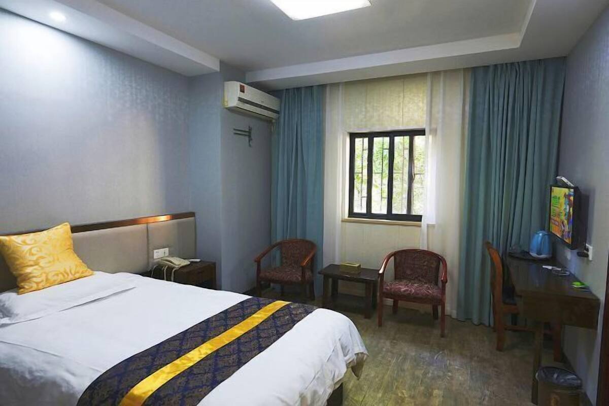 A hotel room with a bed, a desk, two chairs, a window with curtains, and an air conditioning unit.