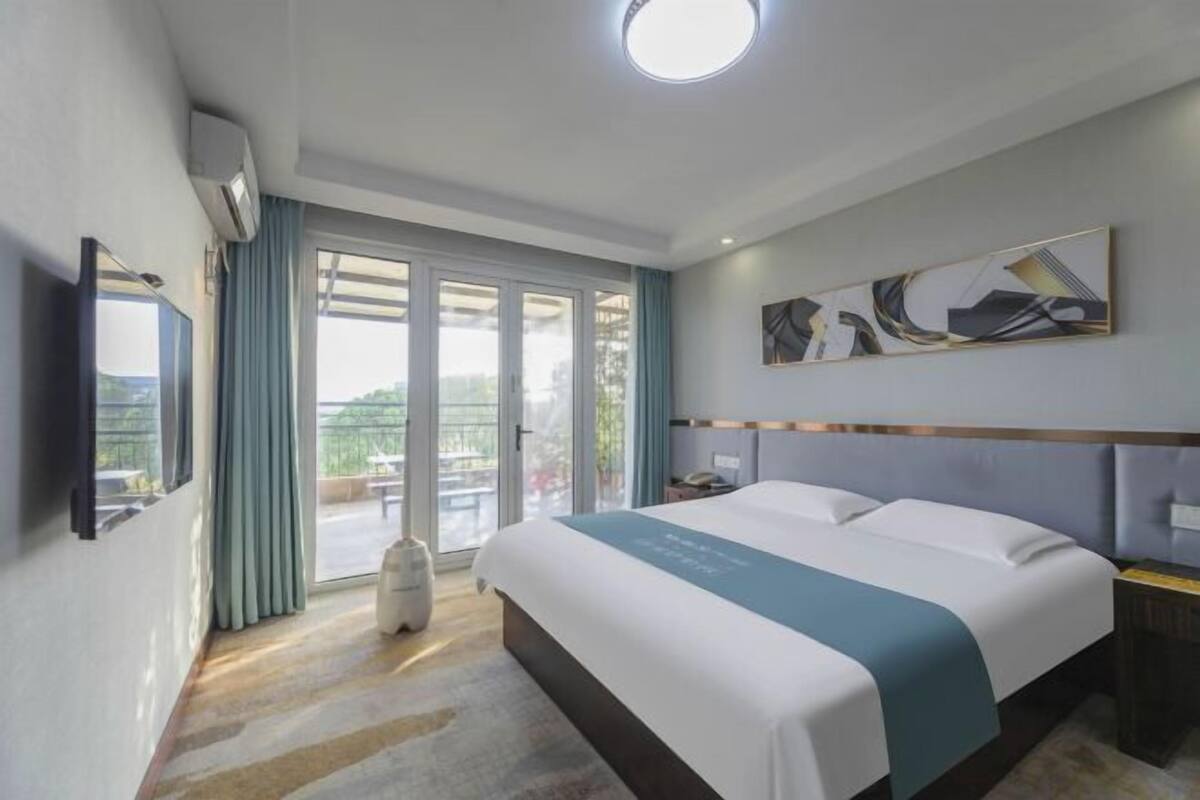 A hotel room with a bed, a flat-screen TV, a large window with curtains, and a ceiling light.