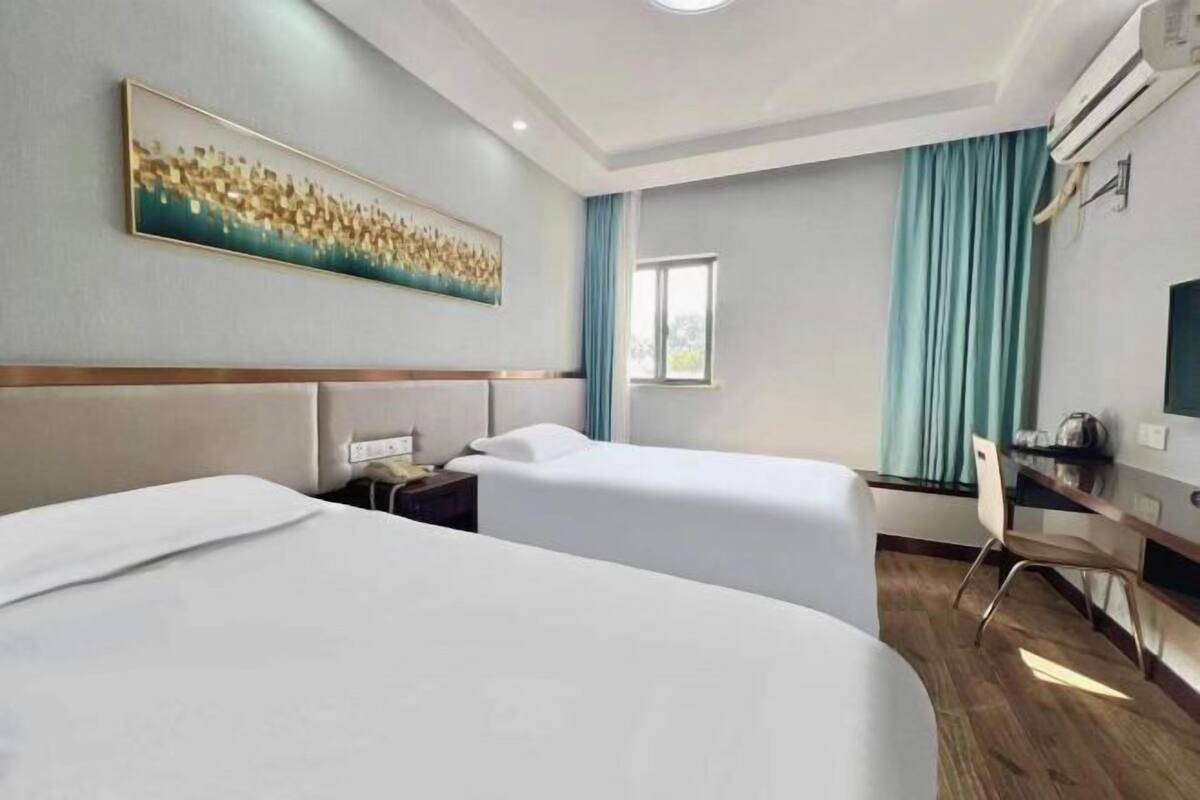 A hotel room with two beds, a desk, a chair, a window with curtains, and an air conditioning unit.