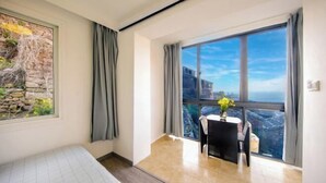 A room with a bed, a table with chairs, a view of the city through large windows, and a stone wall with greenery. - Hongfu Inn (Zhoushan)