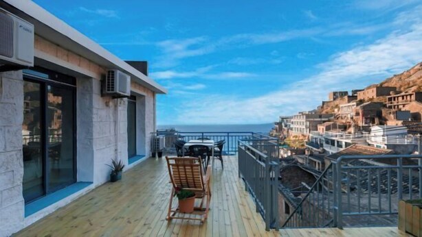 A balcony with a wooden floor, a chair, and a table, overlooking a coastal town with buildings and a clear sky. - Hongfu Inn (Zhoushan)