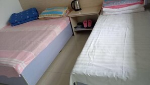 Ein Zimmer mit zwei Betten, eines mit einer rosa-weißen Streifenauflage und das andere mit einer schlichten weißen Auflage. Ein Regal mit Wasserkocher und Hausschuhen, sowie ein kleines Regal mit gefalteter Kleidung.
