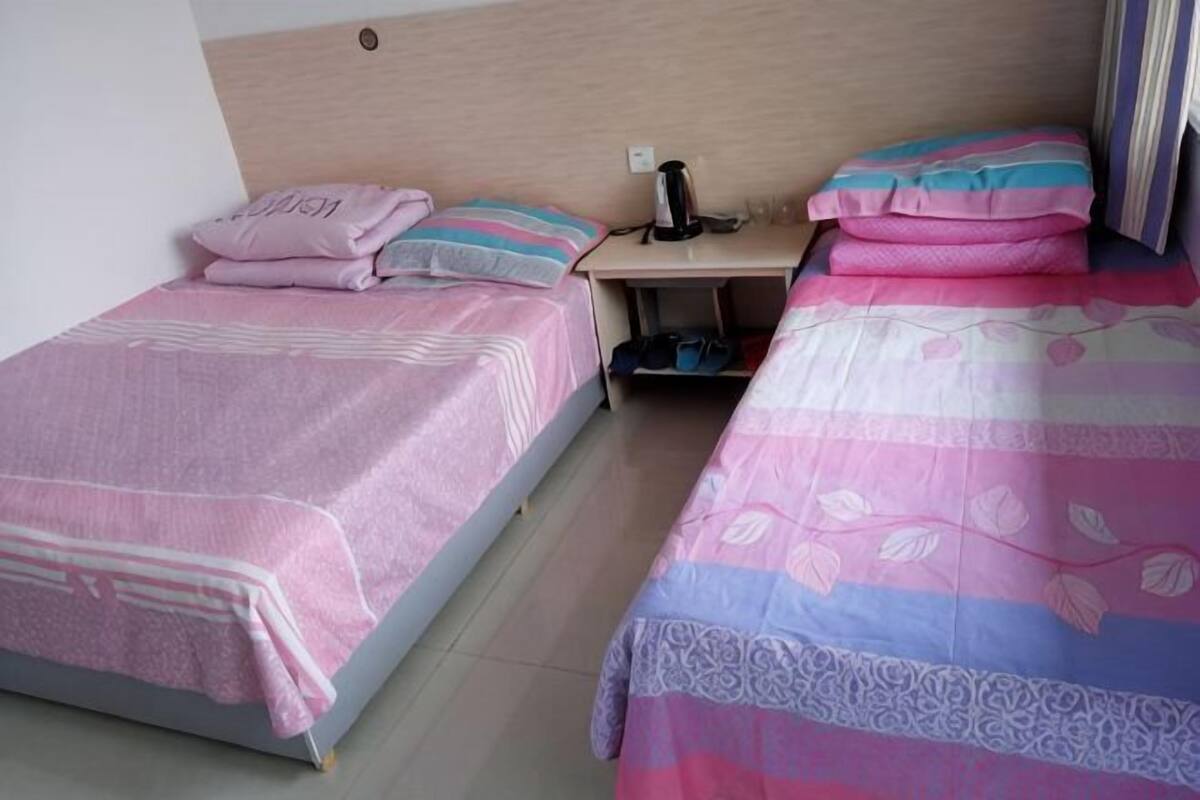 Dos camas individuales con ropa de cama floral en tonos rosa y azul, un cabecero de madera y una mesita de noche con un objeto negro.