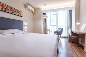Habitación de hotel con cama, escritorio, silla y ventanas amplias.