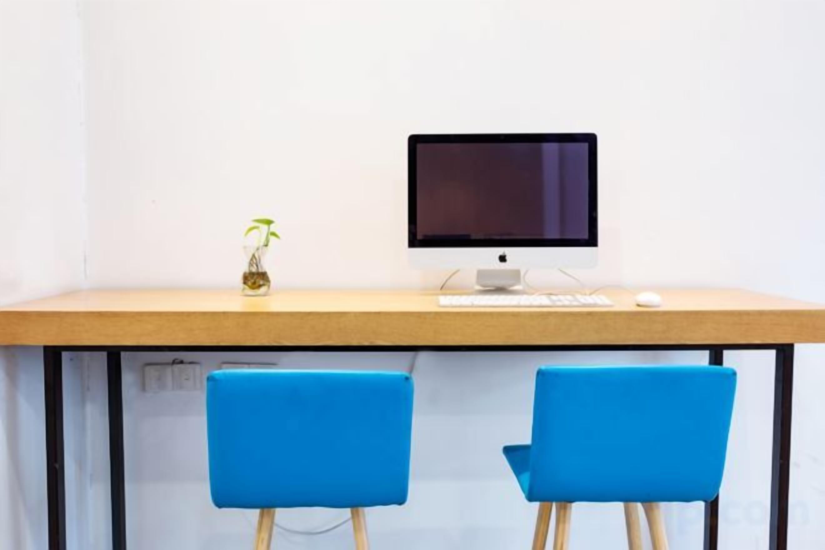 Un'area di lavoro con un monitor per computer, tastiera e mouse su una scrivania in legno. Ci sono due sedie blu e una piccola pianta sulla scrivania.