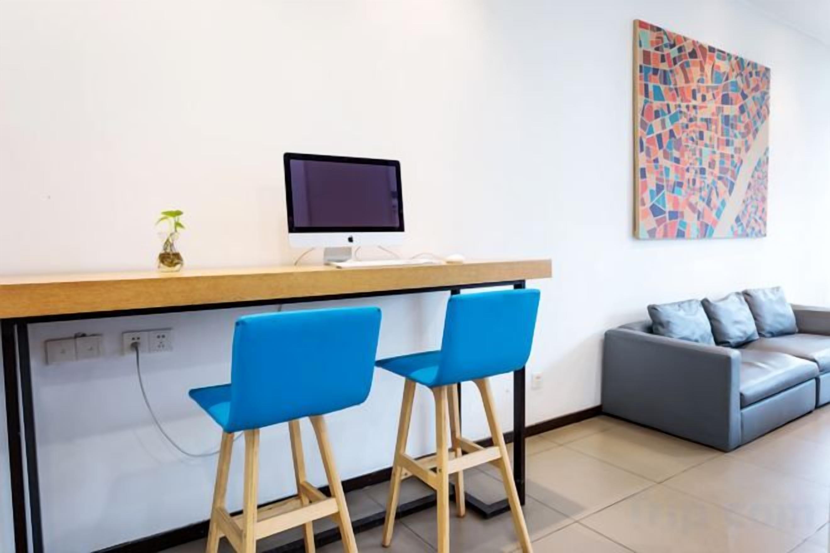 Un espace d’accueil comprenant un bureau, un ordinateur, deux chaises bleues, un canapé gris et un tableau abstrait aux couleurs vives accroché au mur.