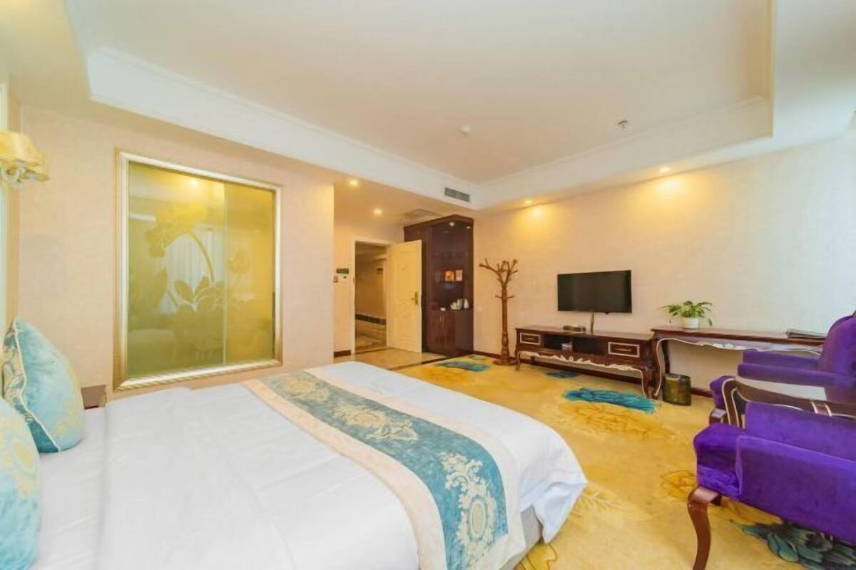 A hotel room with a large bed, a desk, a television, and purple chairs.