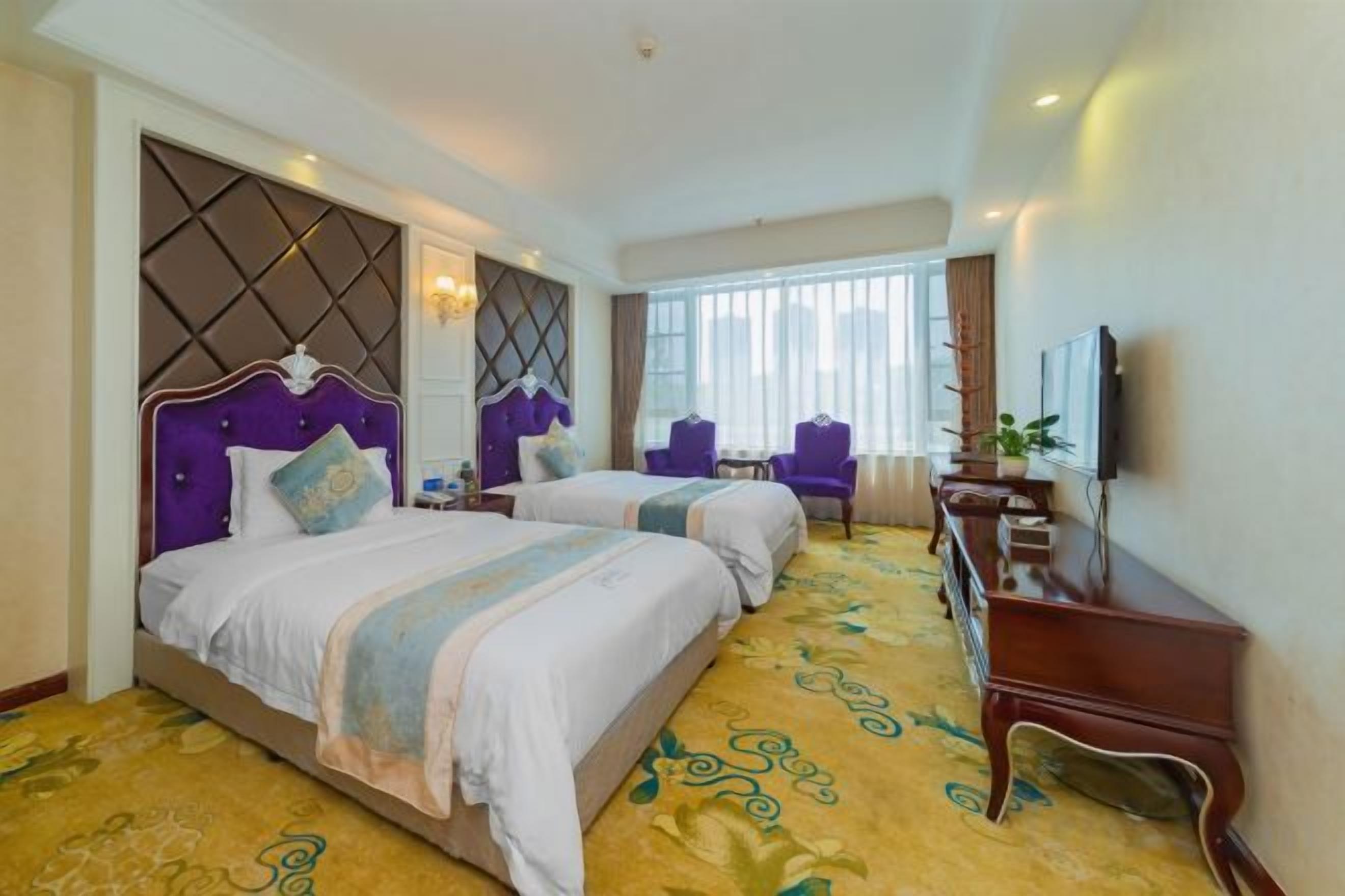 A hotel room with two beds, a desk, a chair, a TV, and a large window.