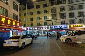 Una strada con auto parcheggiate sul lato, edifici con insegne e alcuni passanti.