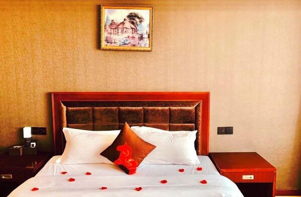 A hotel room with a wooden headboard, a bed with white bedding and rose petals, a nightstand with a lamp, and a framed picture on the wall. - Junyi Chain Hotel (Sichuan Media College) (Chengdu)