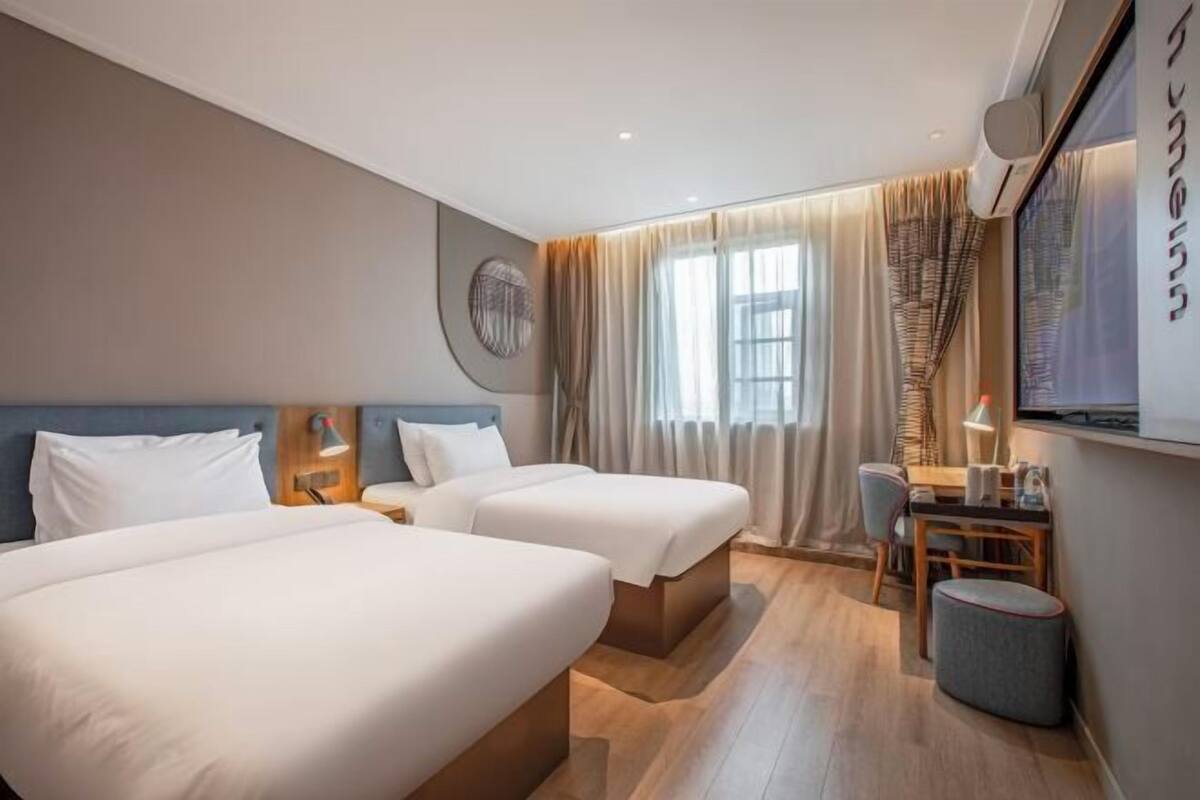 A hotel room with two beds, a desk, a chair, a lamp, and a window with curtains.