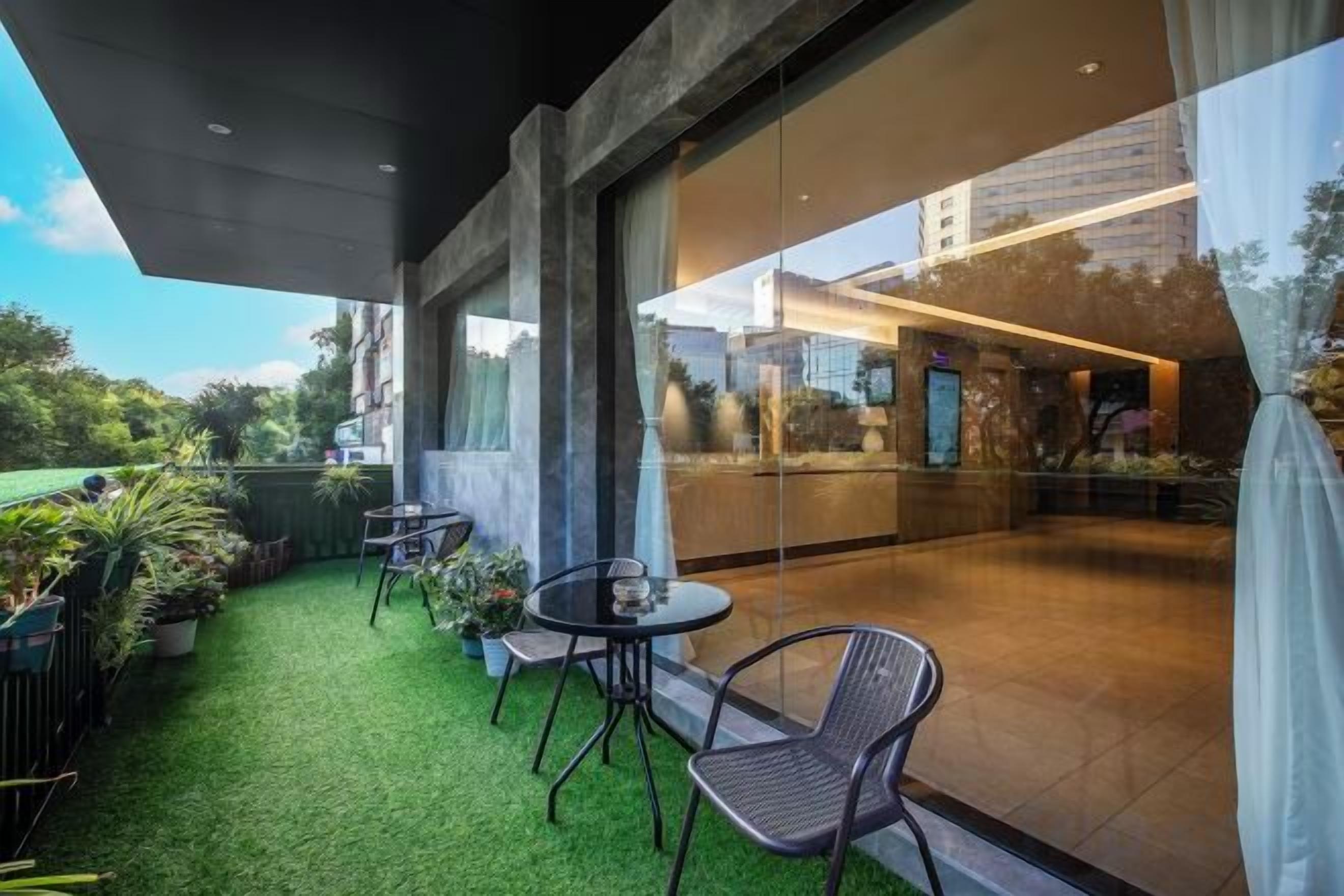 Un área de estar al aire libre con mesas y sillas, césped artificial verde y vista a un edificio a través de un muro de cristal.