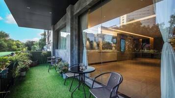 Un área de estar al aire libre con mesas y sillas, césped artificial verde y vista a un edificio a través de un muro de cristal.