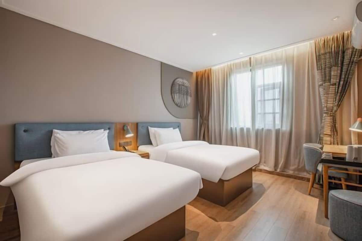 A hotel room with two beds, a desk, a chair, a lamp, and a window with curtains.